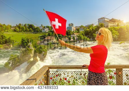 Woman With Swiss Flag On Top Of Swiss Waterfall Rhine Falls In Switzerland At Sunset. The Most Power