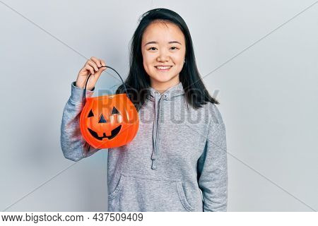 Young chinese girl holding halloween pumpkin basket looking positive and happy standing and smiling with a confident smile showing teeth 
