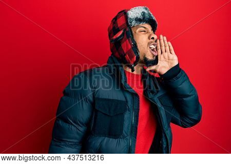 Young african american man with beard wearing winter hat and coat shouting and screaming loud to side with hand on mouth. communication concept. 