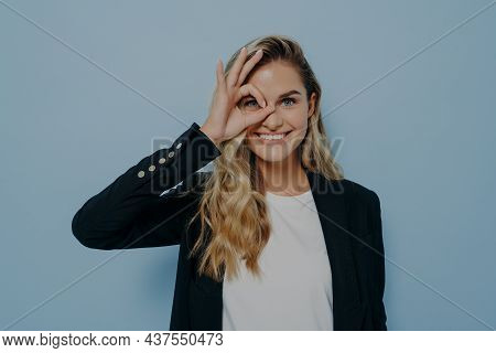 Portrait Of Beautiful Joyful Blonde Caucasian Female Wearing Black Coat Looking Through Okay Sign Ge