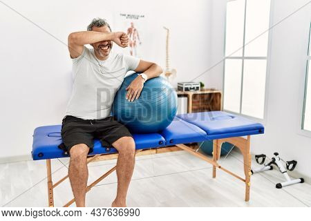 Middle age hispanic man at pain recovery clinic holding pilates ball smiling cheerful playing peek a boo with hands showing face. surprised and exited 