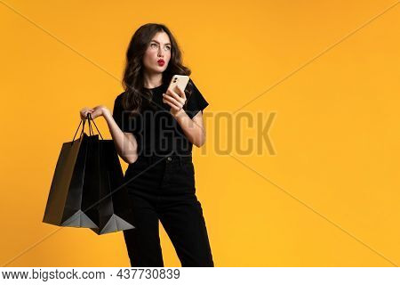 White puzzlement woman posing with shopping bags and cellphone isolated over yellow background
