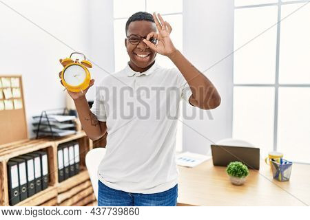 Young african man holding alarm clock at the office smiling happy doing ok sign with hand on eye looking through fingers 