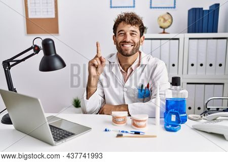 Young hispanic dentist man working at medical clinic showing and pointing up with finger number one while smiling confident and happy. 
