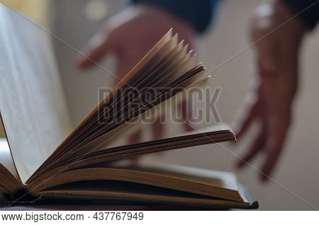 A Defocused Shot Of An Old Open Book And Two Open Male Hands. Palms In The Background. Pages Of Hard