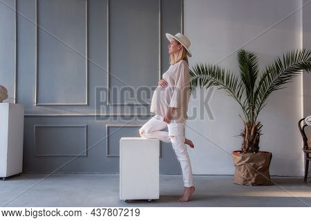 Fashion Pregnant Young Woman In White Hat Leaning Her Leg On A White Cube In A Gray, Stylish Studio