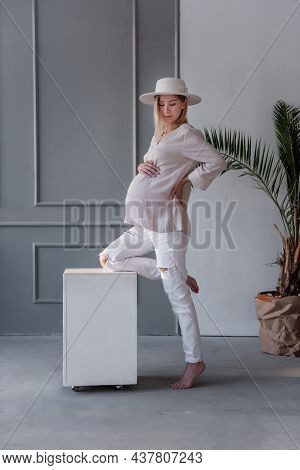 Fashion Pregnant Young Woman In White Hat Leaning Her Leg On A White Cube In A Gray, Stylish Studio