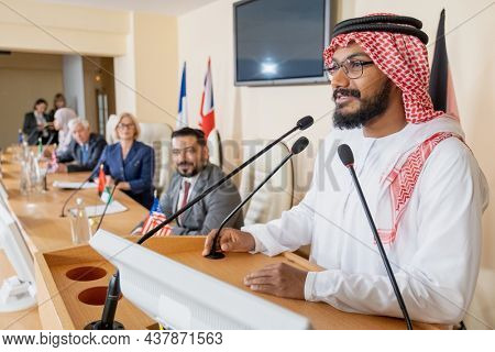 Smiling Muslim politician in ghutrah and agal standing at rostrum podium and speaking in public using mucrophone at international conference
