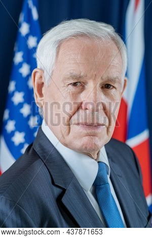 Portrait of serious senior Caucasian congressman in formal suit standing against national flags