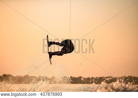 Man Skillfully Making Trick In Jump With Wakeboard On Bright Orange Sky And Sun Background.