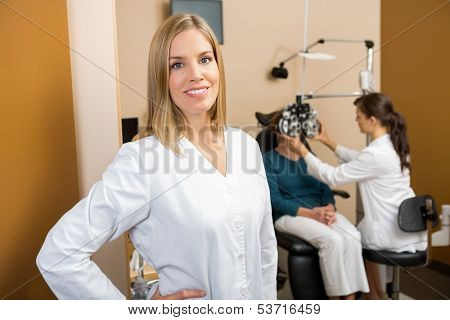 Portrait of mid adult female eye doctor with colleague examining patient in background