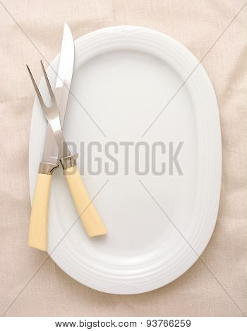 A carving knife and fork on a platter, the platter is oval and white. The carving set has bone handles and the background surface is also off-white. High angle in vertical format.
