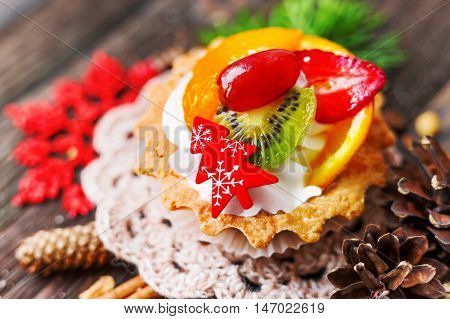 Winter holiday rustic background with fruit tart and spices - cinnamon anise peanuts. Christmas and New Year decorations - red snowflake thuja and pine cones. Tasty dessert with strawberry kiwi orange peach and whipped cream.