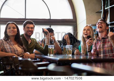 people, leisure, friendship and entertainment concept - happy friends with smartphones drinking beer and watching sport game or football match at bar or pub