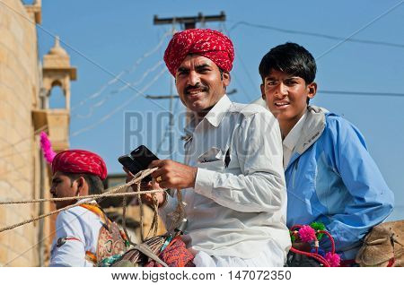 JAISALMER, INDIA - FEB 1, 2015: Two young drovers sitting on a camel with mobile phone in hands during the Desert Festival on February 1, 2015 in Rajasthan. Every winter Jaisalmer takes the Desert Festival
