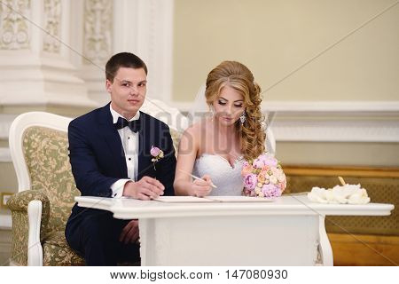 Beauty bride and handsome groom are registering the marriage. Wedding couple is marrying. Beautiful model girl in white dress and in lace veil. Man in suit. Female and male portrait. Cute lady and guy