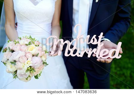 Wedding inscription JUST MARRIED on the nature. Cute couple is holding hands. Beauty bride with groom. Beautiful model girl in white dress. Man in suit. Female and male portrait. Close-up arms