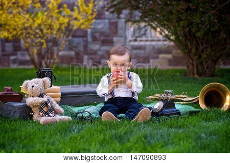Little boy bites apple sitting on a green lawn in a garden. Around the boy are different retro toys