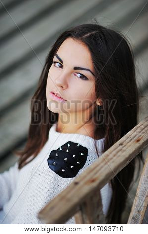 Up portrait of a serious confident beautiful long-haired brunette with piercing brown eyes in a white jacket with spikes on his shoulders on a blurred background with boards sitting in a half-turn.