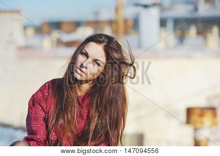 Beautiful thoughtful brunette with long disheveled hair in the wind with blue eyes in a red plaid shirt on a blurred background of city roofs.