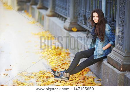 Young Pensive Brunette Girl Sitting On A Concrete Curb Around A Beautiful Iron Fence, Under The Feet