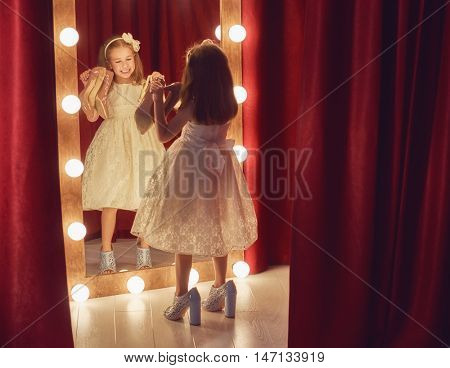 Cute little fashionista. Happy child girl try on outfits and mom's shoes looking at mirror.