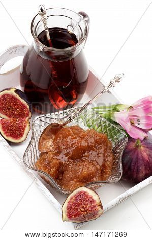 Arrangement of Delicious Homemade Fig Jam in Glass Kremanka with Fresh Fig Fruits Flowers and Jag of Fruit Drink in White Wooden Tray closeup on White background