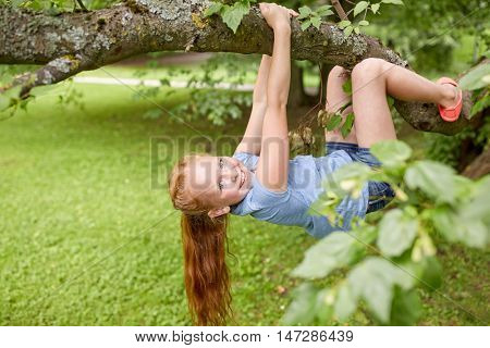 friendship, childhood, leisure and people concept - happy smiling little redhead girl hanging upside down on tree in summer park