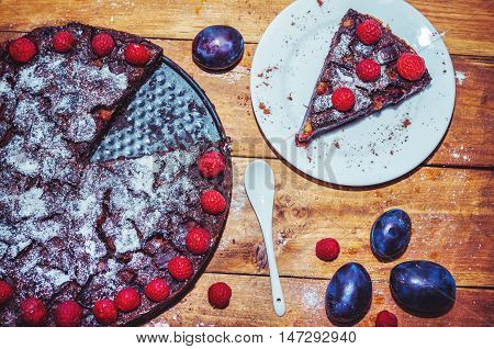 Chocolate clafoutis with plums, decorated by powdered sugar and raspberry in a baking pan, ripe fresh  plums on wooden background