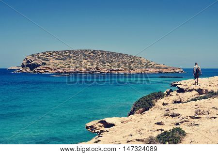 a young tourist man with a camera in his hand walks next to the Mediterranean sea facing the Illa des Bosc island, in Ibiza island, Spain