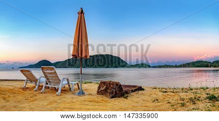 Summer Vacation end concept. Sunset tropical beach with two old chairs and a folded umbrella. Landscape with a quiet sea and the mountain in the distance Koh Chang Thailand.