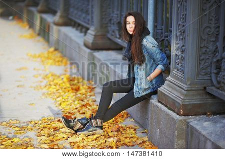 Beautiful dreamy girl in a denim jacket sits on a stone slab near the iron fence of the city park under the feet of autumn maple leaves.