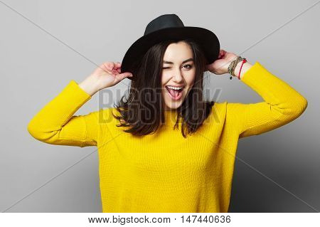 Young beautiful woman in black hat and yellow pullover posing and winking while holding hands on hat. Toned. Grey background