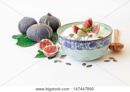 A dish of yogurt with pieces of fig fruit, near scattered sunflower seeds, laid ripe fig fruits on the fig tree leaves on the white background. Yogurt with fig fruit slices. Horizontal. Daylight.