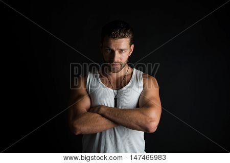 young handsome bearded man, sexy, macho with muscular hands and beard on serious face in white vest posing in studio on black background