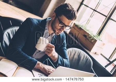 Enjoying fresh coffee while working. Confident young man in smart casual wear working on laptop and holding coffee cup while sitting at his working place in office