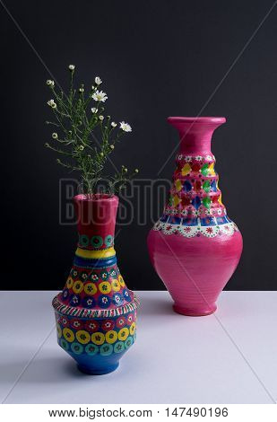 Still life of two artistic painted colorful handcrafted pottery vases little green branches and small white flowers with harsh shadow on white table and black wall