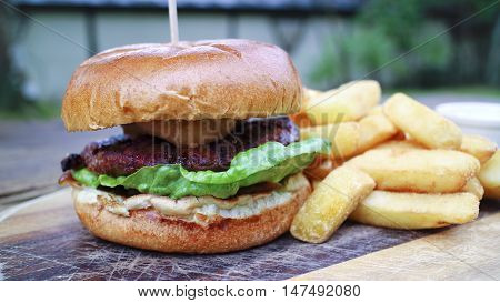 Close up of meat burger with green lettuce relish in a wheat bun offered on wooden board accompanied by chunky potato fries served al fresco in a country pub garden