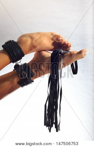 High key photo of sexy female nude legs binded with cuffs holding whip against white background
