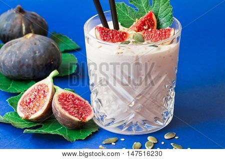 Glass of fig fruit smoothie fig slices straws and near scattered sunflower susami seeds ripe fig fruits on the fig tree leaves on blue background. Fig fruit smoothie glass. Horizontal. Daylight.