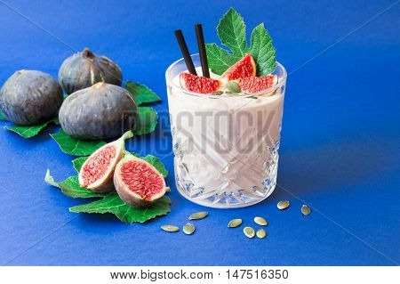 Glass of fig fruit smoothie fig slices straws and near scattered sunflower seeds ripe fig fruits on the fig tree leaves on the blue background. Fig fruit smoothie glass. Horizontal. Daylight.
