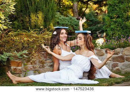 Two cute sisters fooling around with books sitting on the lawn. The girls put books on their heads. They represent themselves Greek nymphs.