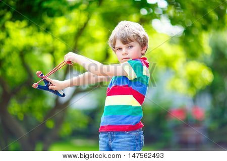 Funny little kid boy shooting wooden slingshot against green tree background. Child having fun in summer.