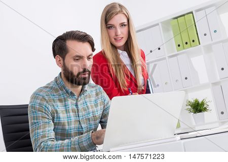 Partners and cofounders of start up at their workplace. Man is working at laptop. Woman is standing by his side and looking to camera. Concept of successful startup