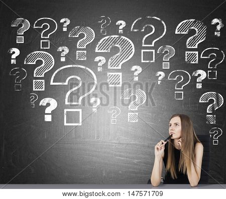 Portrait of businesswoman sitting near chalkboard with multiple question mark sketches. Concept of questions in education