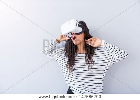 Find new reality. Cheerful delighted woman smiling and using virtual reality device while standing isolated on grey background
