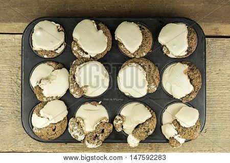 Top down view of one dozen freshly baked home made muffins with brown frosting inside muffin tin on wooden table