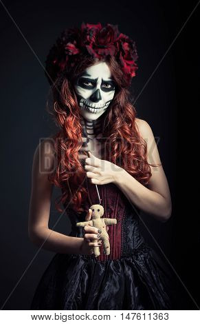 Young voodoo witch with muertos makeup (sugar skull) holds voodoo doll and needle