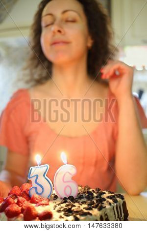 Woman makes a wish before blowing candle in shape of number thirty six on cake, focus on candles