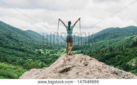 Happy hiker young woman with trekking poles standing on peak of cliff in summer mountains